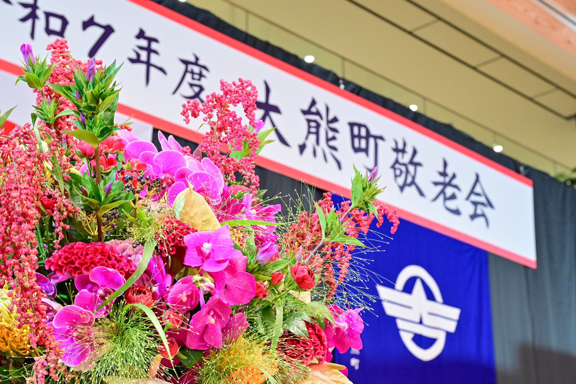 式典会場の壇上には、華やかな花々が飾られました。令和７年度の記念品受賞者は、最高齢（大正12年生・満101歳）1人、米寿53人、しあわせ金婚夫婦13組です。当日は、米寿4人、金婚夫婦8組が出席しました。